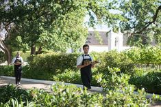 two men in uniforms are walking down the sidewalk with trees and bushes behind them, while one man is holding a clipboard