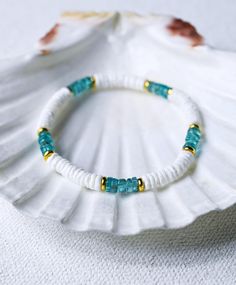 a white bracelet with blue beads and gold accents sits on a shell in the sand