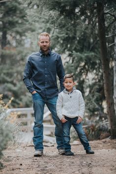 a man standing next to a little boy on top of a dirt road in front of trees