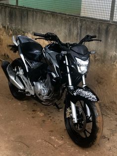 two motorcycles parked next to each other in the dirt near a wall and fenced area