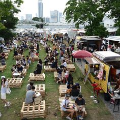 many people are sitting on wooden benches in the grass near food trucks and boats at an outdoor event