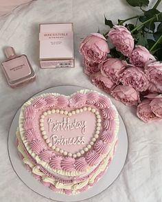 a pink heart shaped cake next to some flowers and perfume bottles on a white table cloth