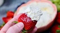 a hand holding a strawberry with whipped cream on it and strawberries in the background