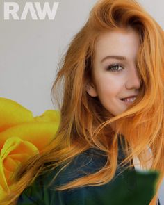 a woman with long red hair holding a yellow rose in her right hand and looking at the camera