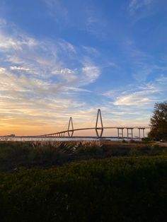 the sun is setting behind a bridge over water with grass and bushes in front of it