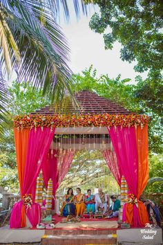 a group of people sitting on top of a lush green field under a pink canopy