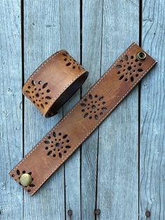 two brown leather wristbands with holes in them on a wooden surface, one has a metal clasp and the other has a brass button