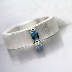 A small blue tourmaline wedding band with a great visual impact. The baguette cut gemstone is set in a yellow gold bar setting and sits atop a wide textured silver band. A minimalistic design with a gorgeous color combination. I fabricated this ring in my studio in Tucson. Size 6 or 52 or 16.5 or L 1/2 (cannot be sized) nangijalajewelry.etsy.com One Of A Kind ( I have more blue tourmalines) Ring Width: 6mm Ring Thickness: 2mm << Blue Tourmaline Specs >> Cut: Baguette Size: 5x2.5mm Weight: 0.20ct Affordable Modern Rectangular Rings, Blue Baguette Cut Ring Fine Jewelry, Timeless Blue Baguette Cut Jewelry, Blue Baguette Cut Topaz Ring Gift, Handmade Rectangular Blue Ring, Blue Aquamarine Baguette-cut Rings, Diamond Band Engagement Ring, Baguette Wedding Band, Wide Silver Ring