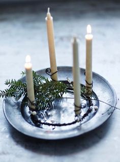 four candles are placed in a metal bowl with greenery on it and one candle is lit