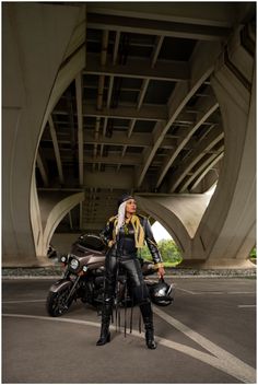 a woman standing next to a motorcycle in the street under a bridge with her hair blowing back