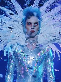 a man with blue hair and white feathers on his head wearing a silver outfit while walking down a runway