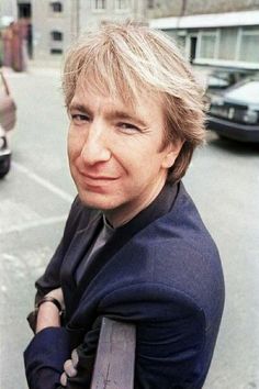 a man with blonde hair and blue shirt leaning on a metal pole in front of a parking lot