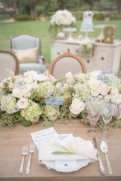 the table is set with white and blue flowers, greenery, and silverware