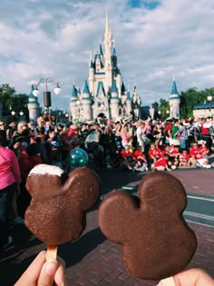 two mickey mouse pops are being held up in front of a castle with people walking by