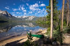 a green canoe sitting on the shore of a lake surrounded by trees and mountains under a blue sky with white clouds