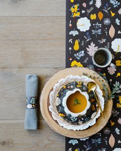 a table with a plate, bowl and spoon on it next to a napkin that has a flower design