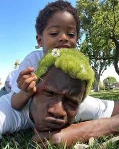 a man and child laying in the grass with green hair on their heads, one is holding a flower