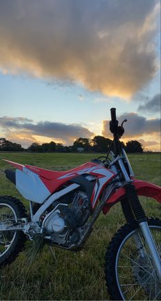 a red and white dirt bike parked on top of a lush green field under a cloudy sky