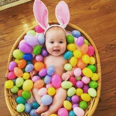 a baby wearing bunny ears in a basket full of candy eggs on the floor,