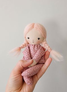 a hand holding a doll with pink hair and white polka dots on the body, in front of a gray background