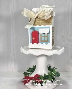 a small house on top of a white cake plate with holly wreath around the base