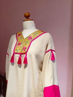 Embrace the charm of traditional Tunisian attire with our "Mony Kamraya" Jebba, a stunning blend of cultural heritage and modern flair. Crafted in a soft, textured fabric, this elegant piece features a rich cream base highlighted with vibrant pink accents. The intricate, hand-embroidered yoke showcases a lively floral pattern adorned with gold thread, while playful tassels add a touch of whimsy. Designed in a one-size-fits-all standard size, this Jebba is perfect for any occasion, ensuring you s Traditional Kaftan With Tassels, Festive Cream Kaftan With Resham Embroidery, Festive Multicolor Kurta With Tassels, Traditional Multicolor Kurta With Tassels, Traditional Pink Kaftan With Traditional Drape, Cream Resham Embroidery Kaftan For Festive Occasions, Cream Kaftan With Resham Embroidery For Festive Occasions, Festive Kurta With Tassels, Festive Bollywood Kurta With Tassels