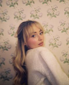 a woman with long blonde hair standing in front of a floral wallpapered background