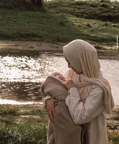 a woman holding a baby in her arms near a body of water with grass and flowers