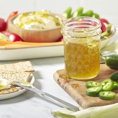 a jar of honey sits on a cutting board next to crackers and green peppers