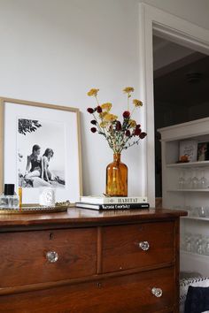 a vase with flowers on top of a wooden dresser next to two pictures and wine glasses