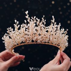 two hands holding a tiara in front of a black background with gold and white beads