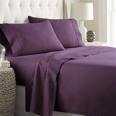 a bed with purple sheets and pillows in a room next to a lamp on a table