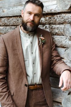 a man with a beard wearing a brown suit and flower boutonniere standing in front of a wooden wall