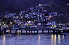 a pier at night with lights reflecting off the water and houses on top of a hill in the background