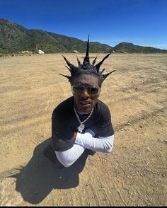 a man with spiked hair sitting in the middle of a dirt field wearing sunglasses and a black t - shirt