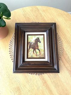 a painting of a horse in a brown frame on a wooden table next to a potted plant