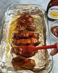 a person holding a brush over some chicken wings on tin foil with sauce in the background