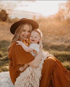 a woman holding a baby in her arms while sitting on the ground with grass and trees behind her