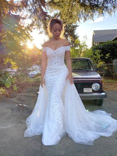 a woman standing in front of a car wearing a wedding dress with off the shoulder sleeves