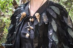 a woman wearing a black dress with feathers and skulls on it's shoulders is standing in the woods