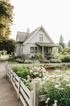 a white house surrounded by flowers and greenery with a wooden fence in the foreground