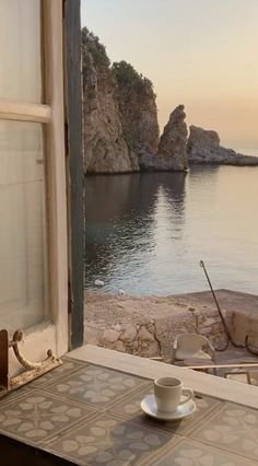 a cup and saucer sitting on top of a table near a window overlooking the ocean