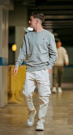 a young man walking down a hallway in white pants and a grey sweatshirt with his hands in his pockets