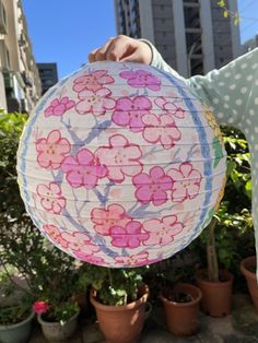 a person holding up a paper lantern with flowers on it in front of some potted plants