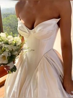 a woman in a white wedding dress holding a bridal bouquet and looking out the window