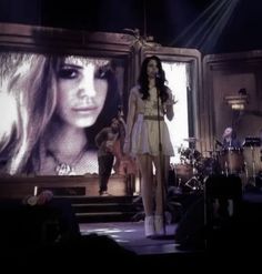 a woman standing on stage in front of a large screen with music playing behind her