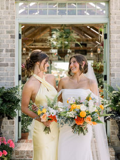 two bridesmaids standing in front of a brick building with flowers and greenery