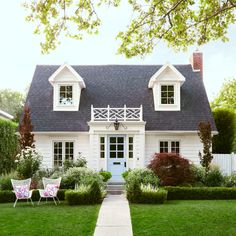 a white house with two lawn chairs in front of it and bushes around the yard