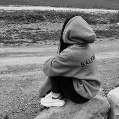 a black and white photo of a person sitting on a rock wearing a hoodie