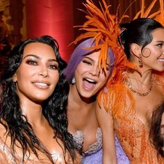 three beautiful women posing for the camera with feathers on their head and one woman in an orange dress